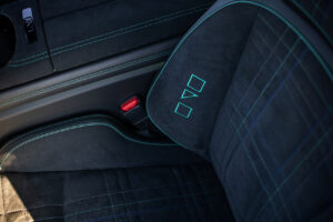 파란색 스티치와 격자무늬 패턴이 있는 검은색 스웨이드 시트, 조명이 있는 시트 조절 버튼, 빨간색 안전벨트 버클이 있는 차량 내부를 클로즈업한 모습입니다.