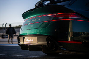 해질녘 레이싱 트랙에 주차된 녹색 제네시스 스포츠카의 후미를 클로즈업하여 흐릿한 배경과 자동차 미등을 비추는 사람 사이를 걷고 있는 모습을 보여줍니다.