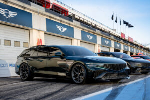 매끄러운 검은색 스포츠 왜건 한 대가 차고 앞 경주로에 주차되어 있고, 그 옆에는 또 다른 스포츠카가 있습니다. 배경은 청명한 하늘과 차고 위의 레이싱 배너로 현대적입니다.