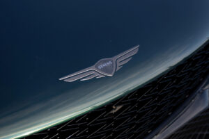 짙은 녹색 제네시스 차량의 전면 후드를 클로즈업하여 검은색 메쉬 그릴 위에 제네시스 날개 엠블럼이 보이고 매끄러운 표면에 햇빛이 반사되고 있습니다.